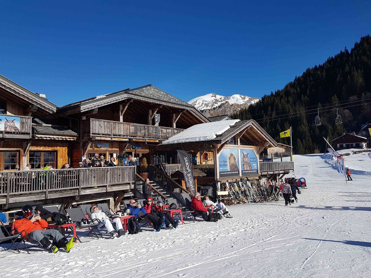 Vue extérieure ensoleillée du restaurant Les Aigles du Léman à Morzine avec du public qui profite du soleil sur les transats.